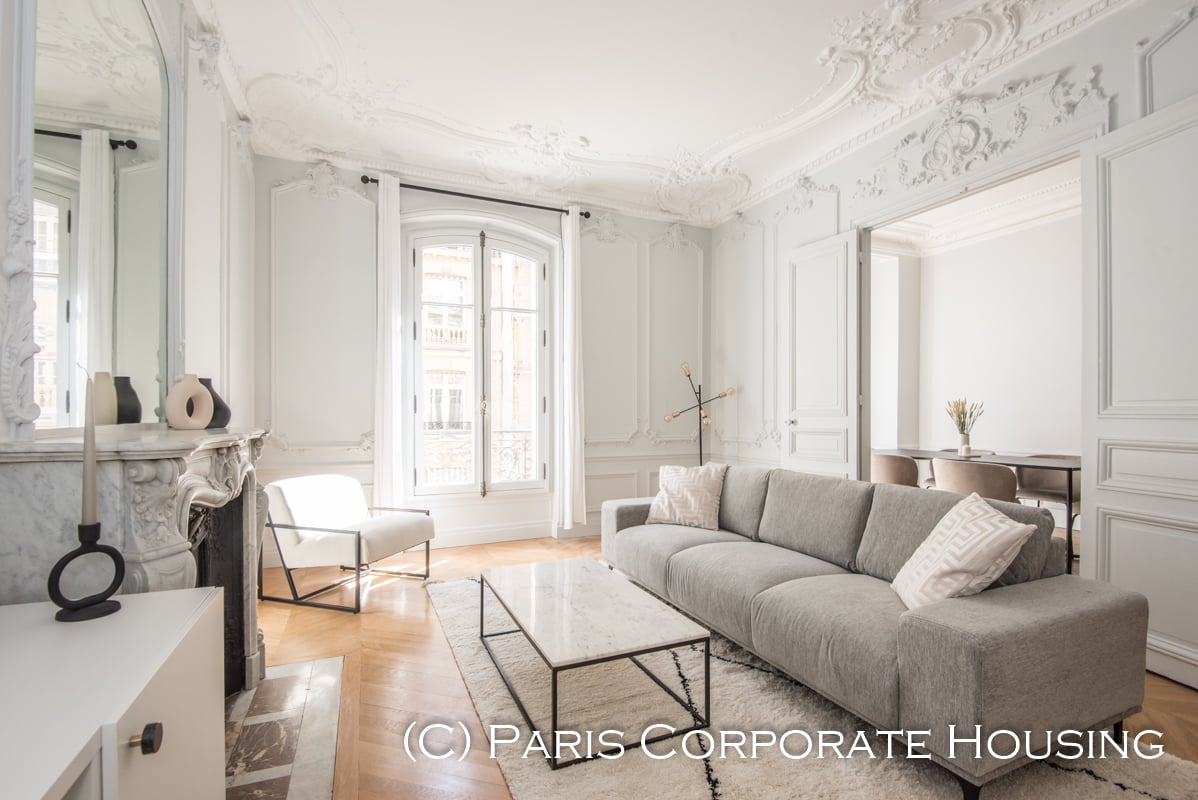 Elegant Haussmann apartment with ornate ceiling details, abundant natural light, marble fireplace, and sophisticated contemporary furnishings in prestigious Parisian location.